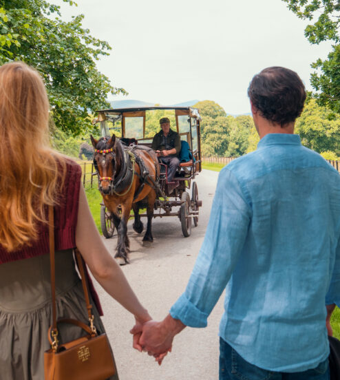Couple holding hands walking through Killarney National Park with a jaunting car