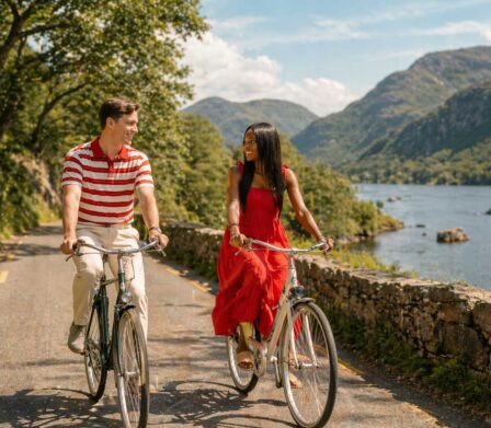 Couple cycling by a scenic lakeside with mountains, blue sky, lush greenery; ideal for romantic getaways and nature lovers.