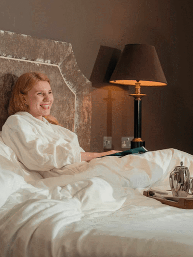 Guest relaxes in cosy hotel room with plush bed, elegant lamp, and breakfast tray; enjoy comfort and warmth.