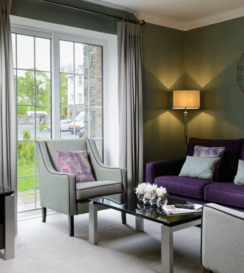 Elegant hotel lounge with plush purple sofa, modern décor, flat-screen TV, and large window view of lush greenery.