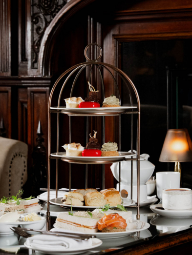Afternoon tea tiered stand with sandwiches, scones and pastries by a roaring fireplace in a cosy hotel lounge