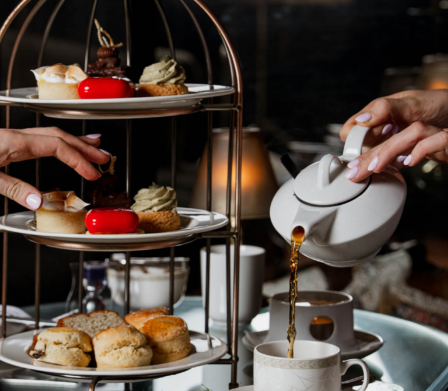 Elegante merienda con una bandeja de varios pisos con scones, pasteles y sándwiches, mientras se sirve el té en una fina taza de porcelana en un acogedor salón de hotel.