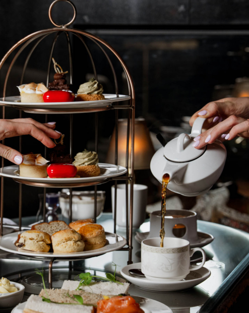 Elegant afternoon tea with tiered stand of scones, pastries and finger sandwiches while tea is poured into a fine china cup in a cosy hotel lounge