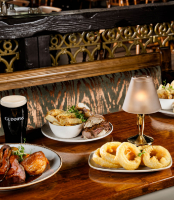 Selection of pub dishes including roast meat, sandwiches, chips and onion rings on a wooden table.