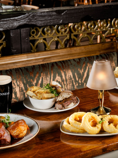 Selection of pub dishes including roast meat, sandwiches, chips and onion rings on a wooden table.