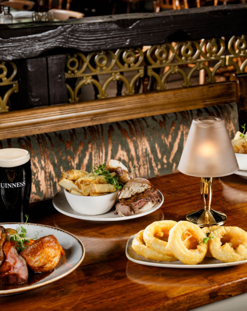 Selection of pub dishes including roast meat, sandwiches, chips and onion rings on a wooden table.