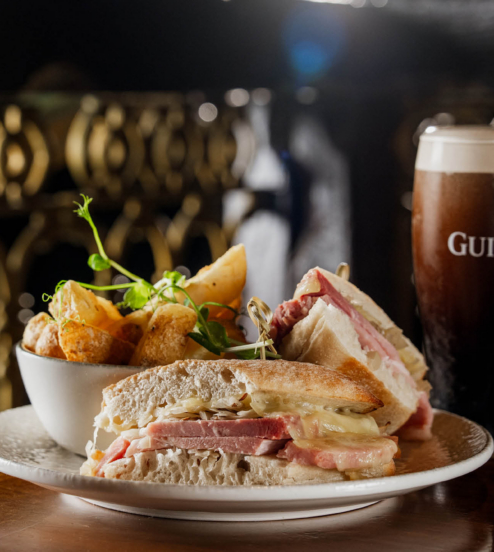 Warm toasted sandwich with melted cheese and ham, chunky potato wedges in a bowl and a pint of Guinness on a wooden pub table, cosy bar setting